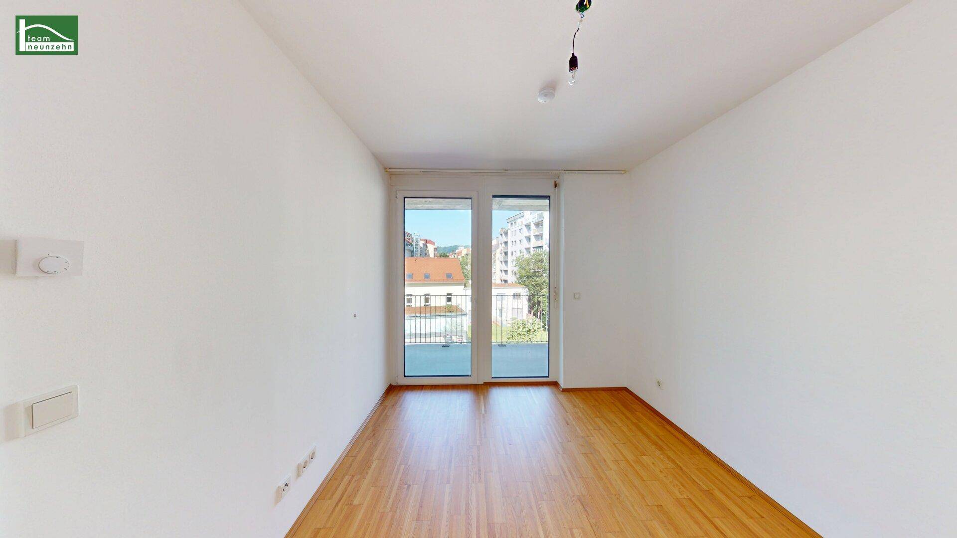 Leeres Zimmer mit Holzboden und großen Fenstern, die einen hellen und offenen Eindruck vermitteln.