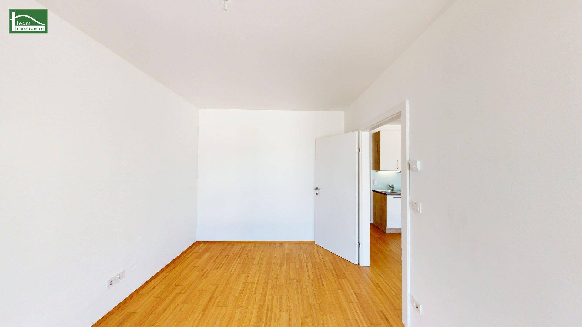 Heller, leerer Raum mit Holzfußboden und offener Tür, die zu einem anderen Bereich führt.