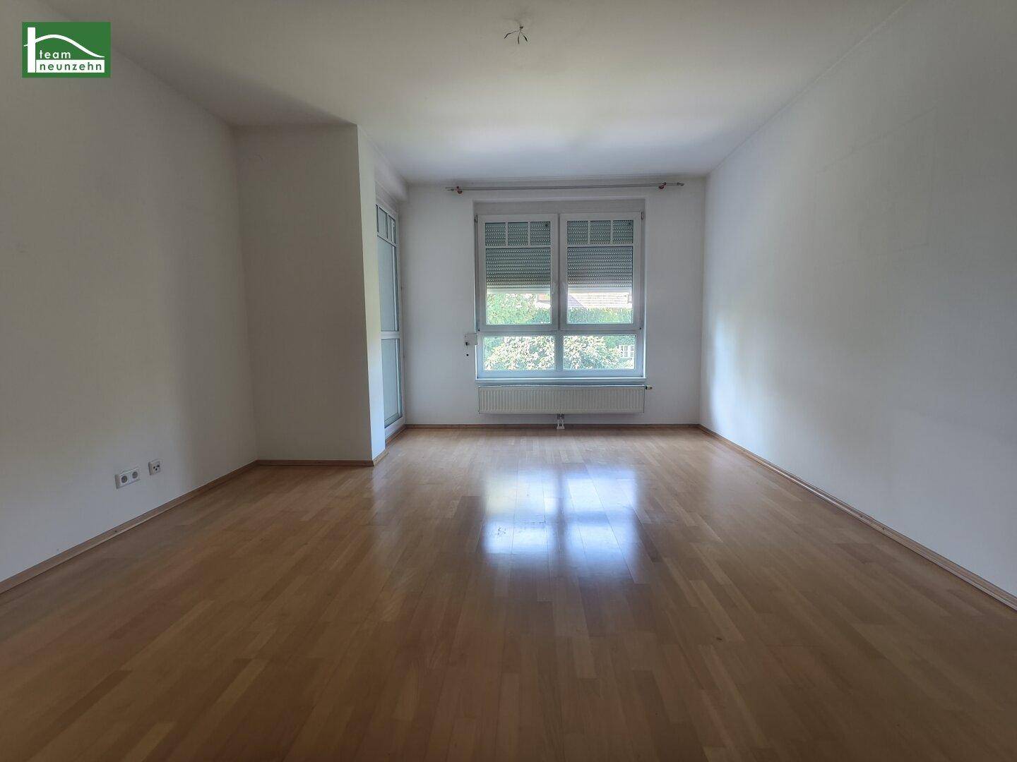 Helles, leeres Zimmer mit Parkettboden und Fenster mit Blick ins Grüne.