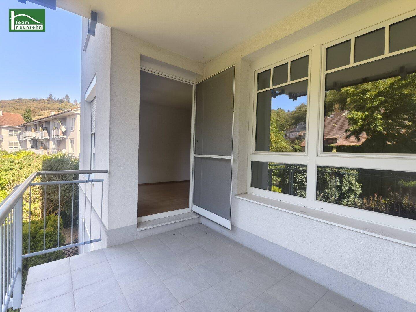 Sonniger Balkon mit Fliesenboden und Zugang zu einem Innenraum, umgeben von viel Grün.