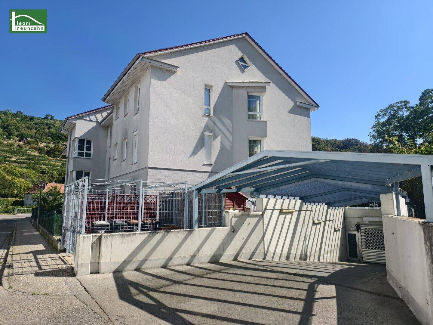Seitenansicht des Gebäudes mit Carport und Zufahrt, umgeben von grüner Landschaft.
