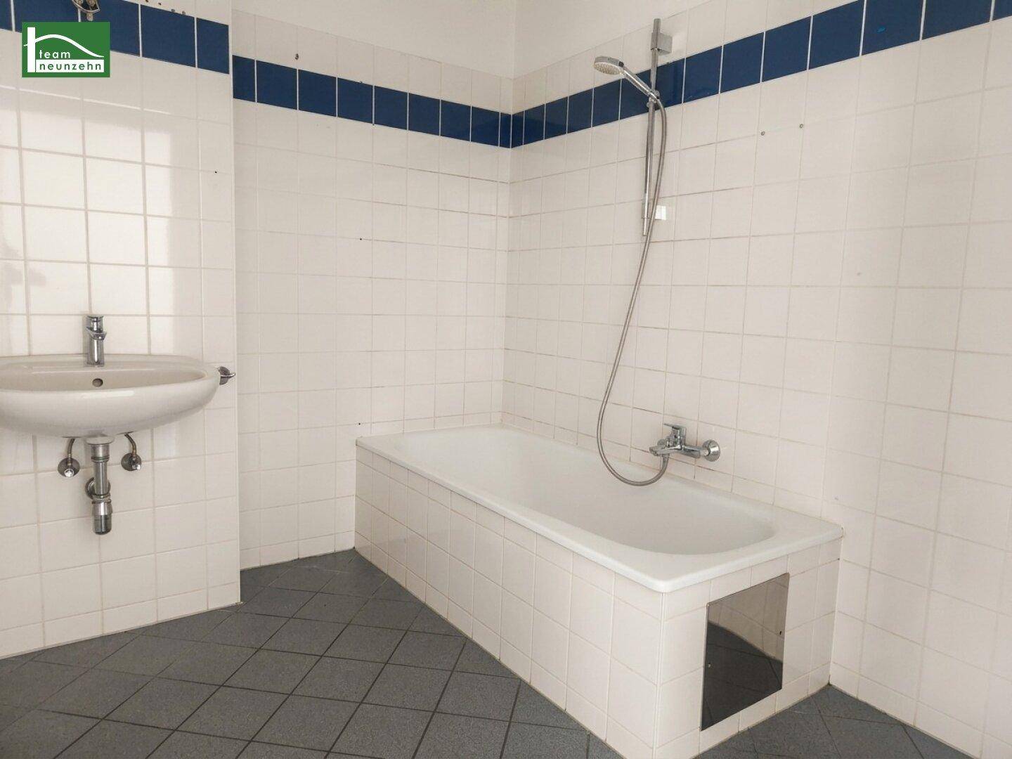 Badezimmer mit Badewanne, Waschbecken und weißen Fliesen mit blauer Akzentlinie.