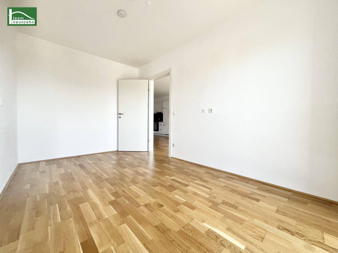 Heller Raum mit Parkettboden und weiß gestrichenen Wänden, offen zu einem weiteren Bereich.
