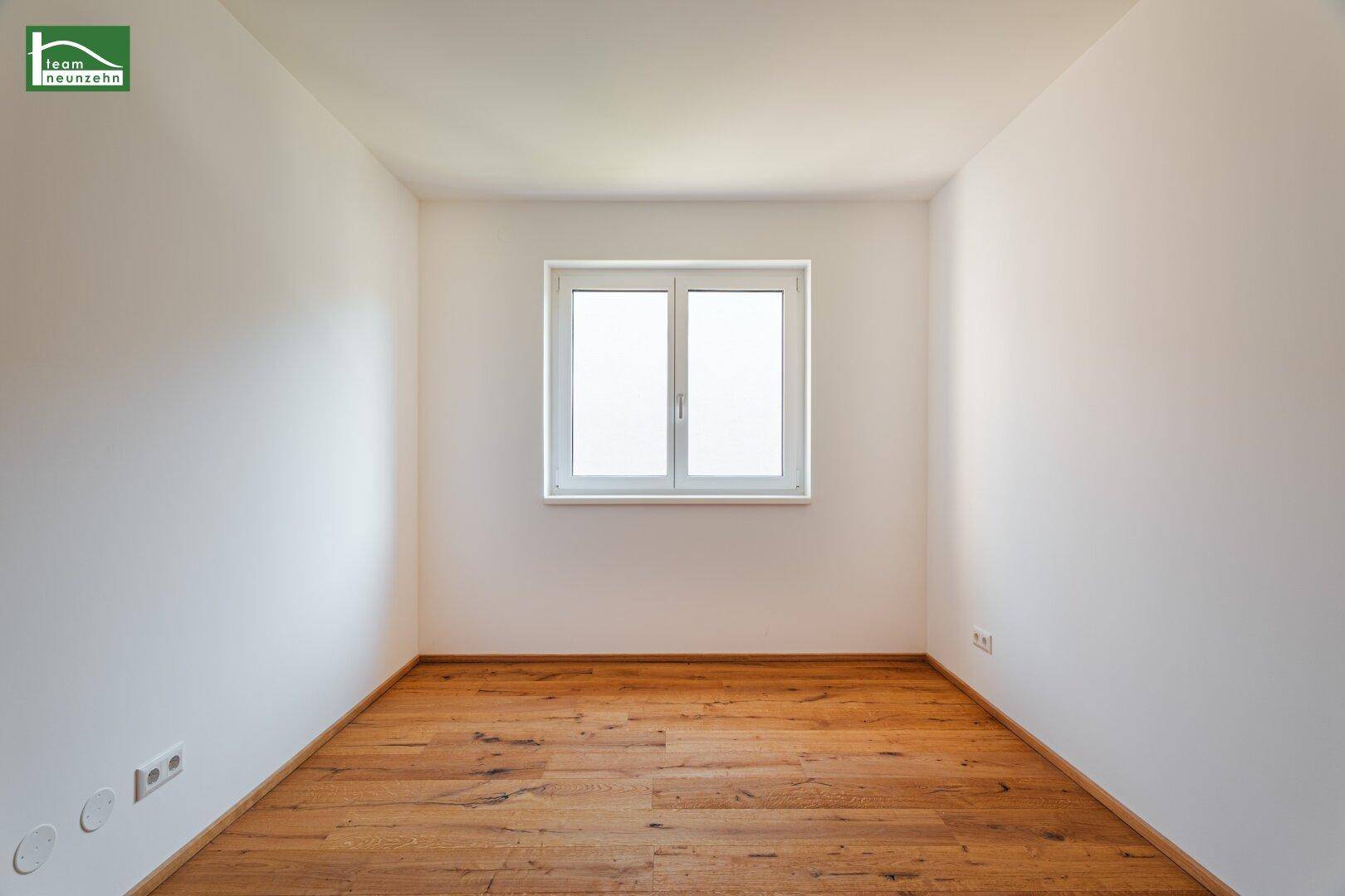 Neutral gestaltetes Zimmer mit Holzboden und einem großen Fenster für viel Tageslicht.