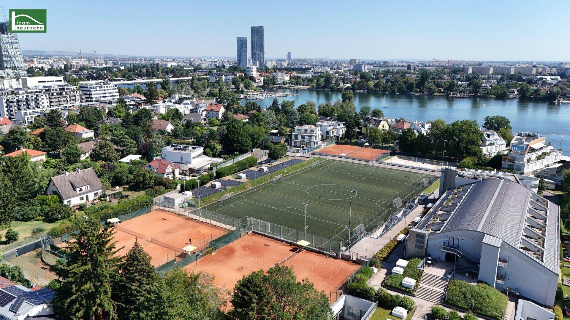 Luftaufnahme mit Sportplätzen, See und städtischer Bebauung in der Ferne.