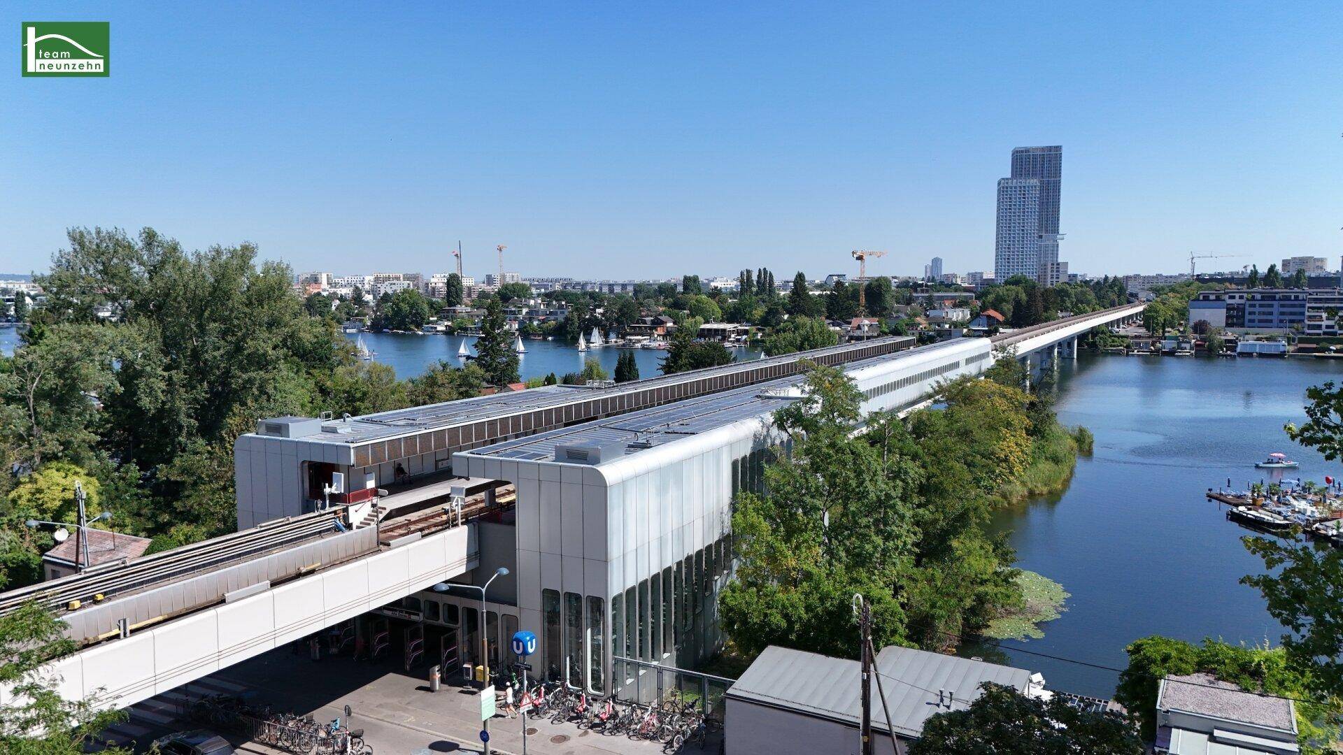 Luftaufnahme einer modernen Bahnstation über dem Wasser mit Stadtkulisse.