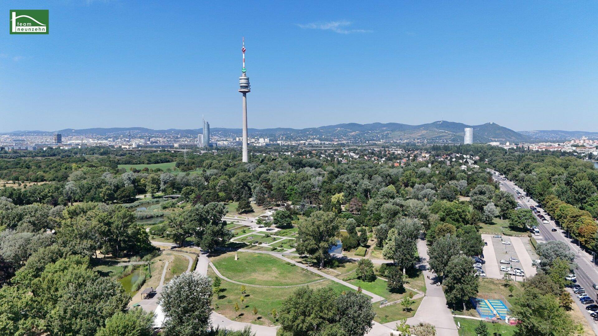 Luftaufnahme eines weitläufigen Parks mit Bäumen, Wegen und einem Fernsehturm am Horizont.