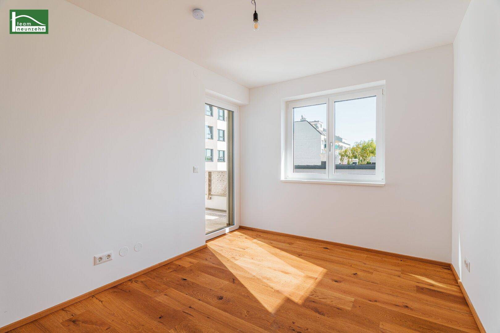 Helles Zimmer mit Holzboden, großen Fenstern und Zugang zu einem Außenbereich.