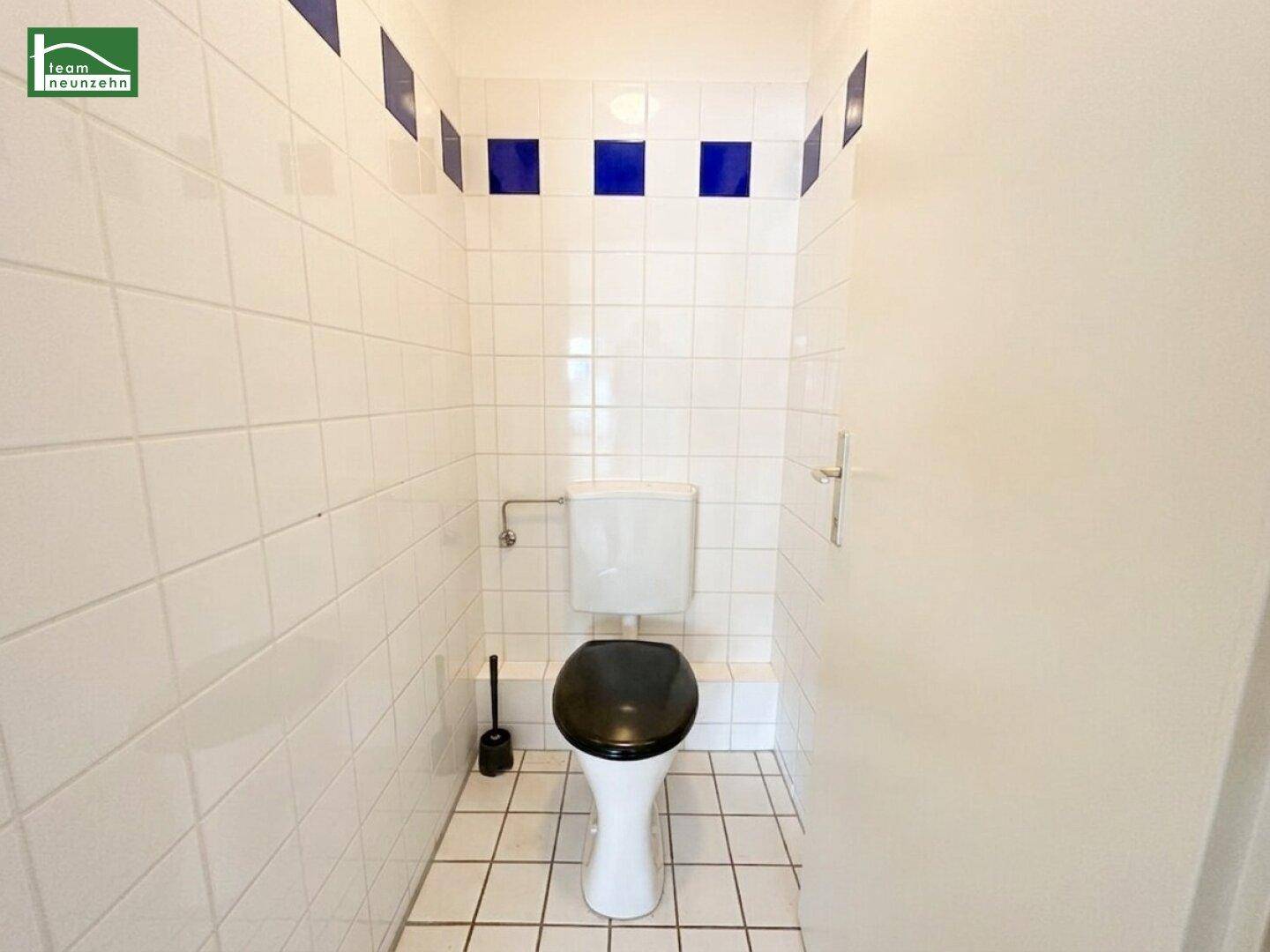 Separate Toilette mit weiß gefliesten Wänden und einem dunklen Toilettensitz.
