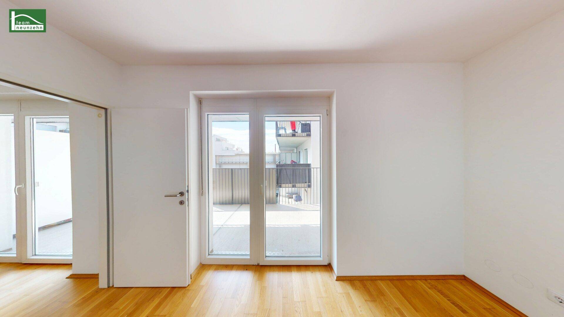 Geräumiges, leeres Zimmer mit Holzboden und Zugang zu einem sonnigen Außenbereich.