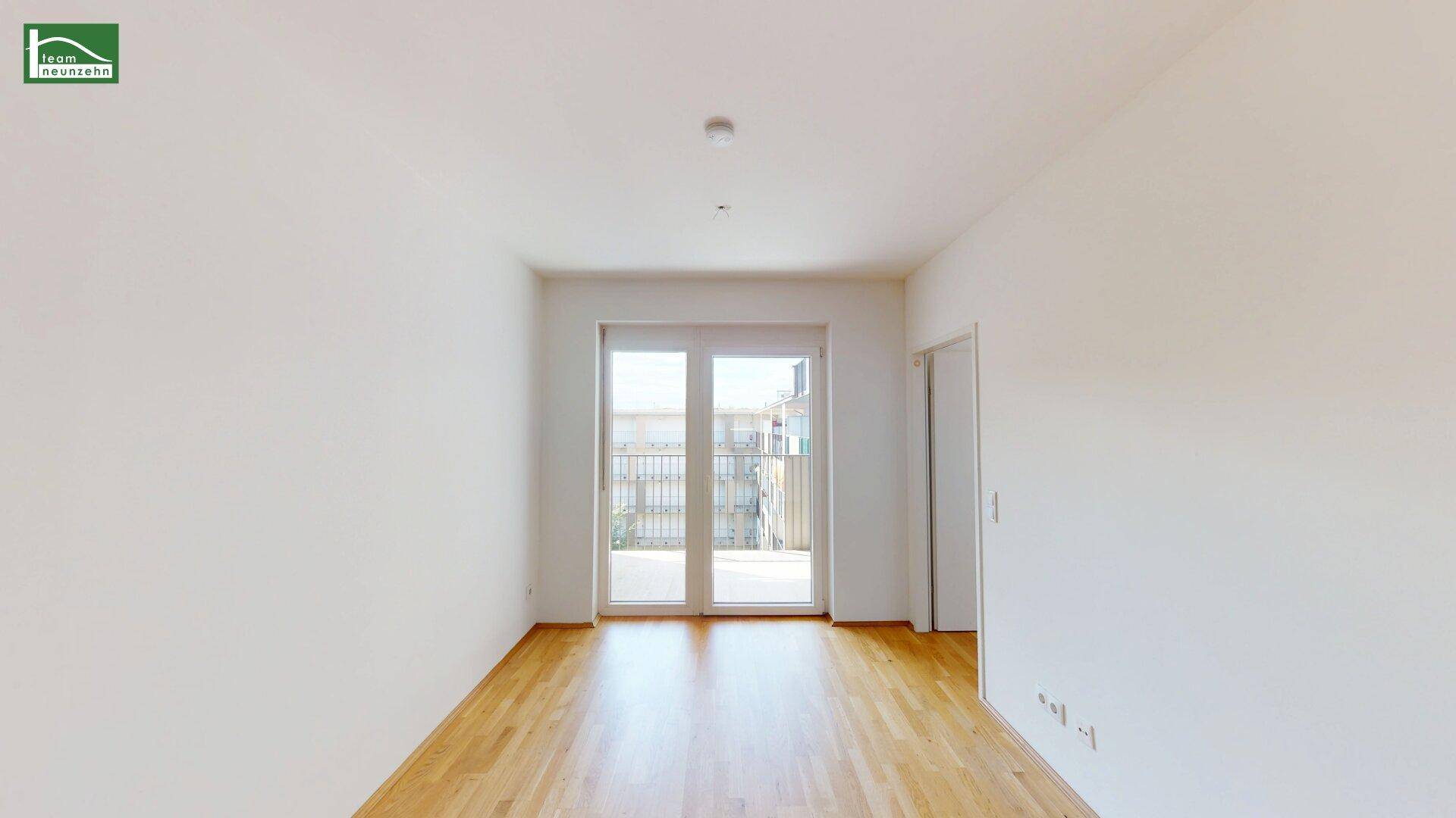 Lichtdurchfluteter Raum mit Holzboden und breiter Glasfront, die zu einem Balkon oder einer Terrasse führt.