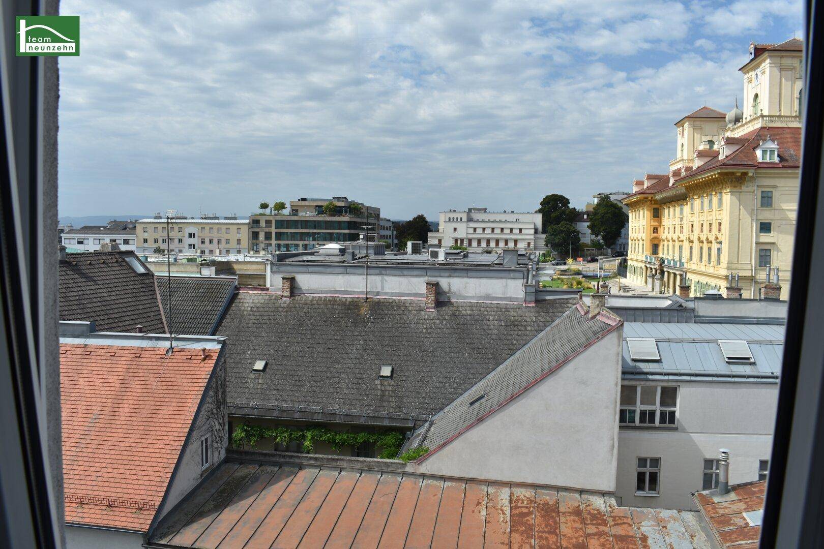 Ausblick aus dem Fenster über die Dächer der Stadt und umliegende Gebäude unter bewölktem Himmel.