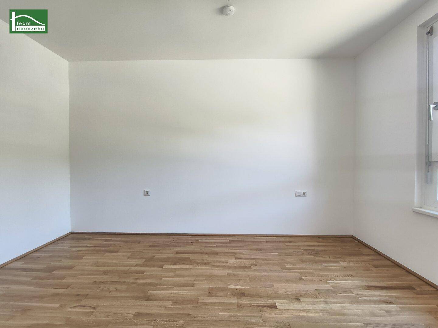 Helles, leeres Zimmer mit Holzboden und einem Fenster, das viel Tageslicht hereinlässt.