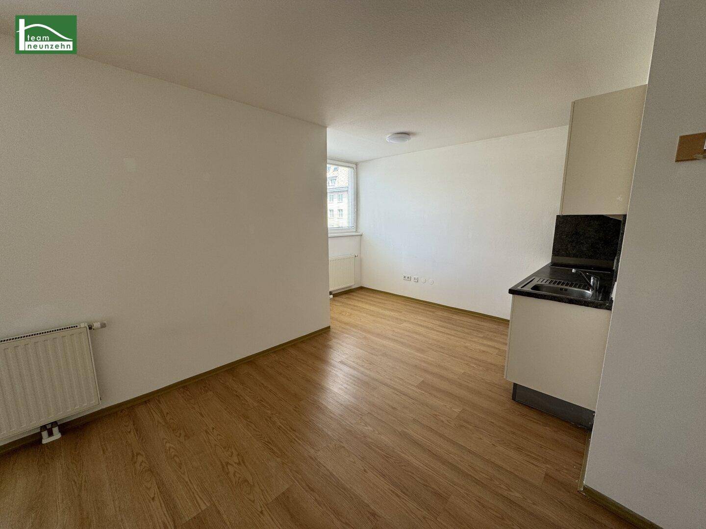 Leerer, heller Raum mit Laminatboden und Heizkörper unter einem Fenster.
