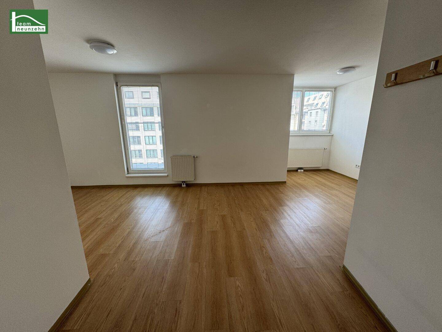 Heller Wohnbereich mit zwei Fenstern, Laminatboden und weiß gestrichenen Wänden.