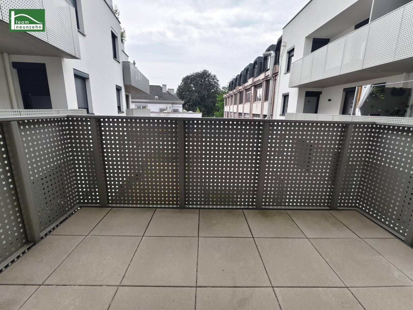 Moderner Balkon mit Fliesenboden und perforierter Metallbrüstung, umgeben von Wohngebäuden.
