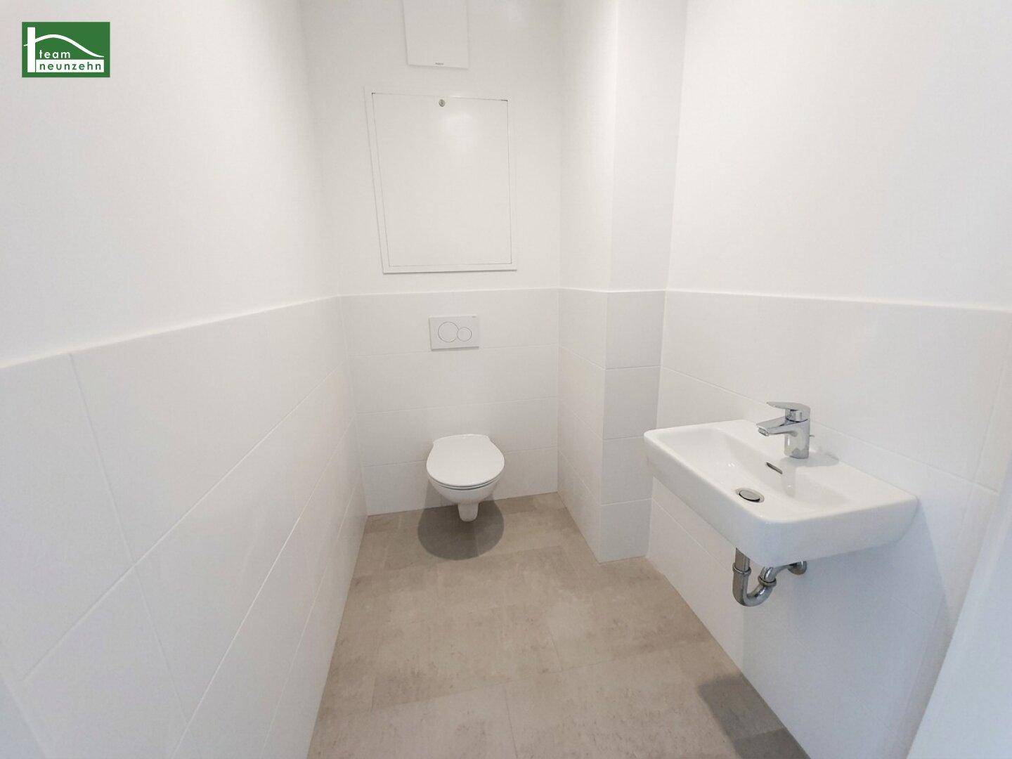 Separates WC mit wandhängender Toilette und kleinem Handwaschbecken, hell gefliest.