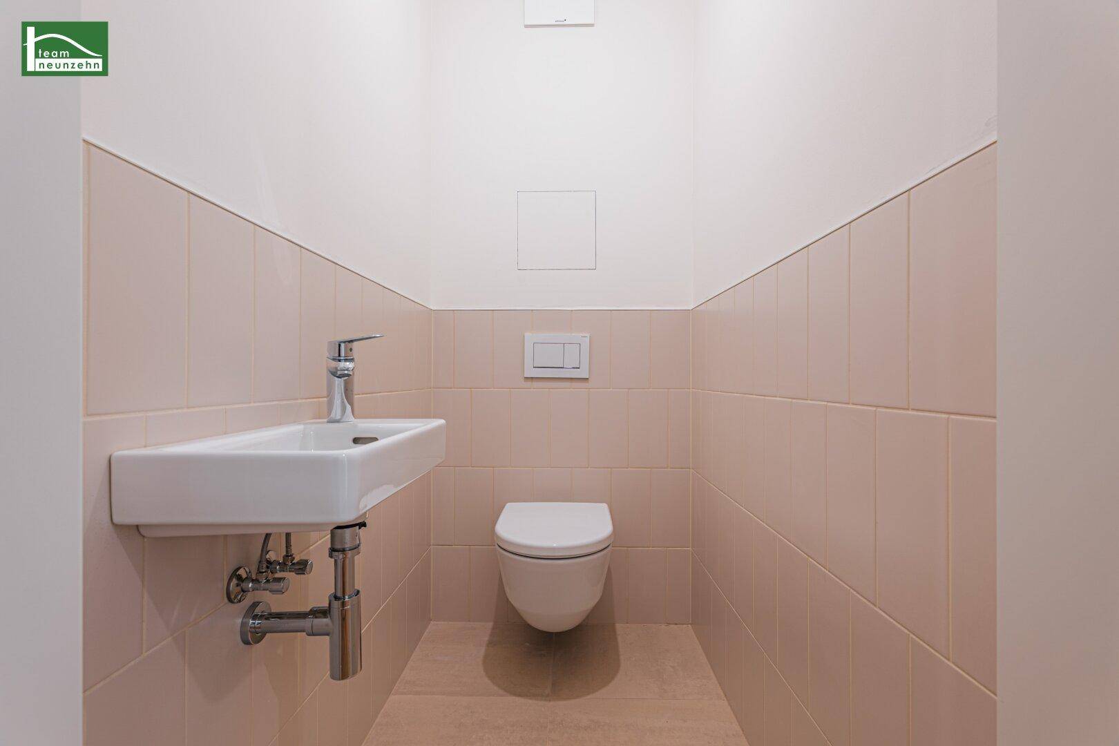 Separates WC mit beigefarbenen Fliesen, wandhängender Toilette und kleinem Handwaschbecken.
