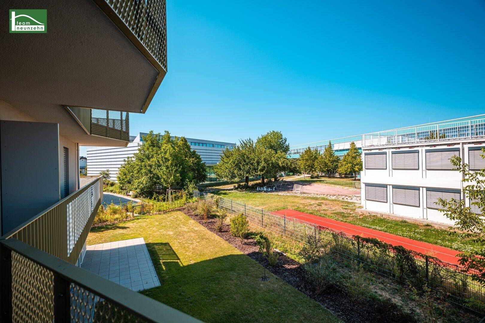 Balkon mit Aussicht auf eine gepflegte Rasenfläche und Sportanlagen in der Ferne an einem sonnigen Tag.