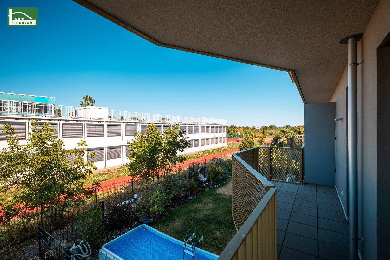 Balkon mit Blick auf einen Gartenbereich, einen kleinen Pool und umliegende Gebäude an einem sonnigen Tag.
