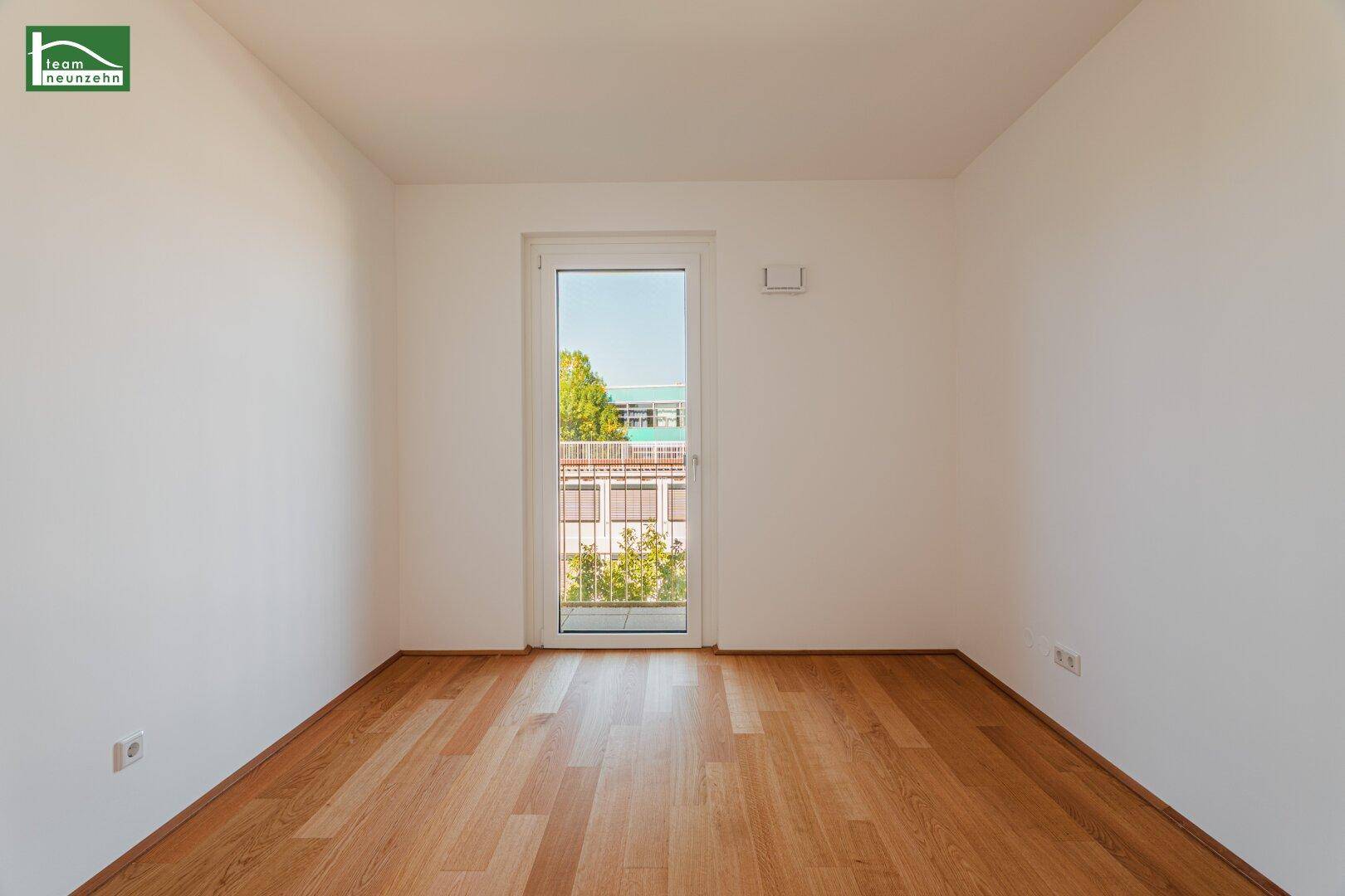 Leerer Raum mit Holzboden und einer Balkontür, die viel natürliches Licht hereinlässt.