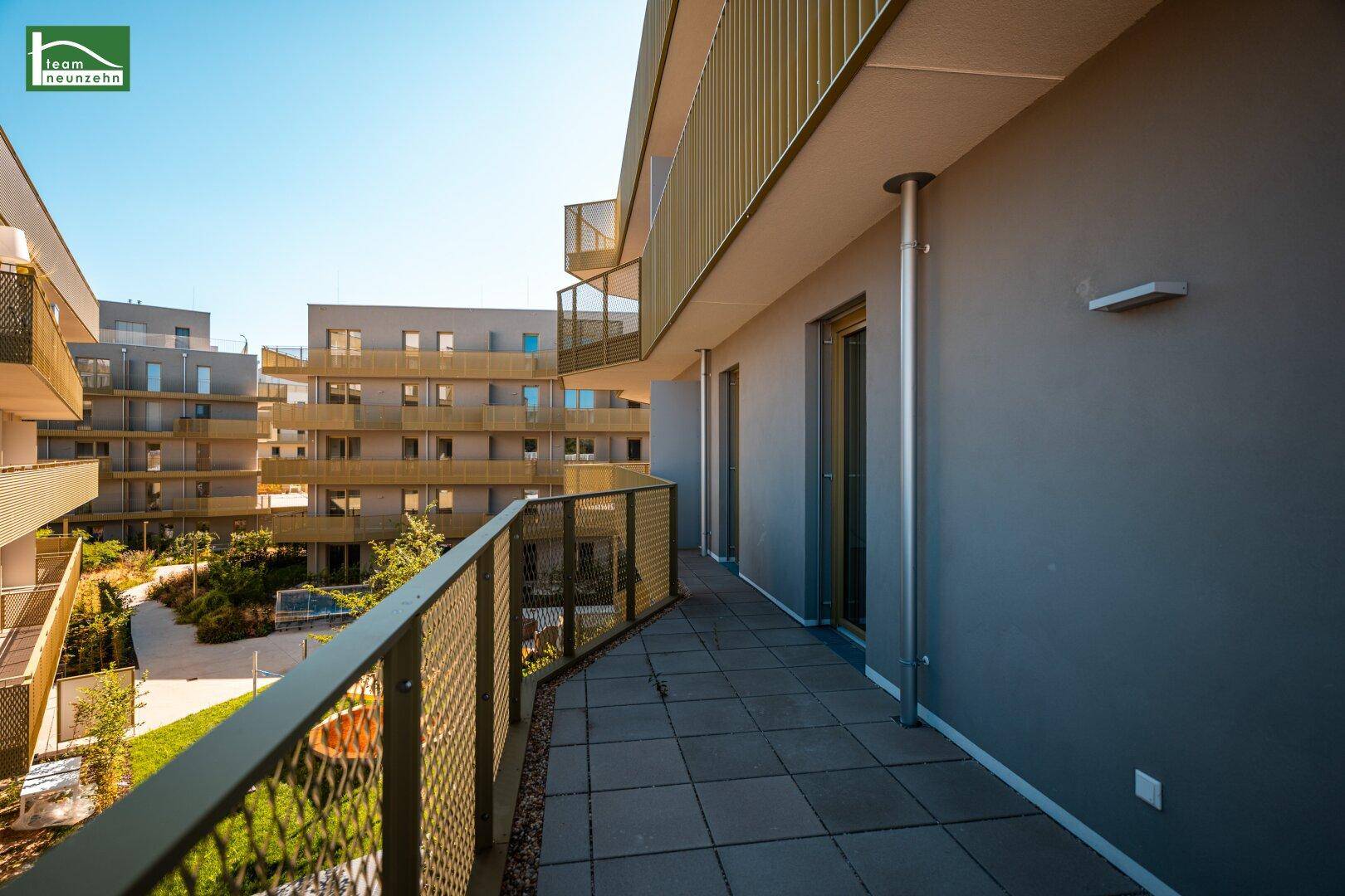 Langer Balkon mit Fliesenboden und Blick auf die moderne Wohnanlage und Grünflächen.