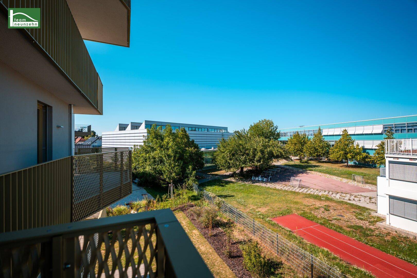 Balkon mit Blick auf eine Grünfläche und Sportanlagen unter blauem Himmel.