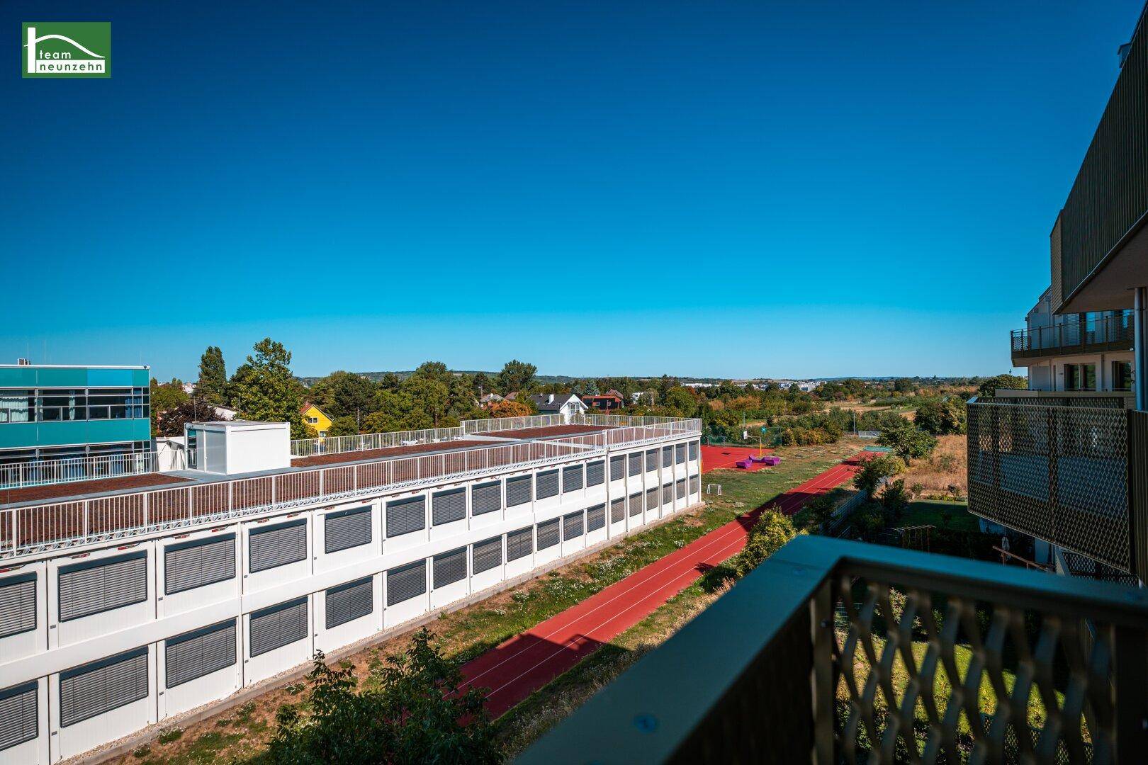 Weitblick vom Balkon über eine Sportanlage und grüne Landschaft unter blauem Himmel.