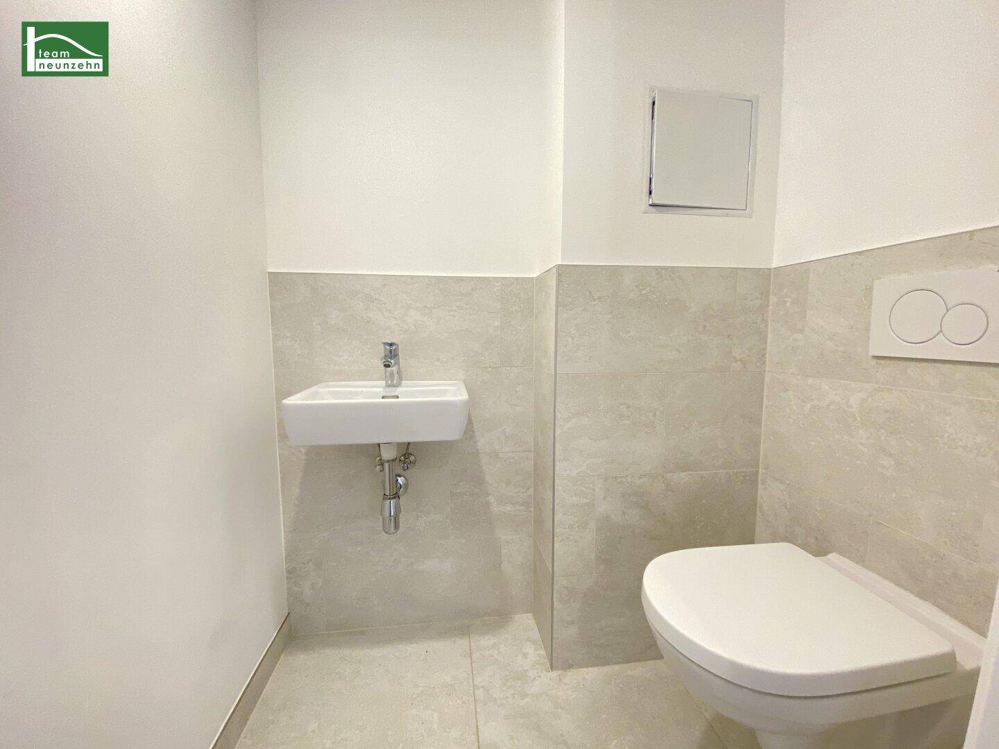 Modernes Gäste-WC mit Wand-WC, kleinem Waschbecken und hellen Fliesen an Wänden und Boden.