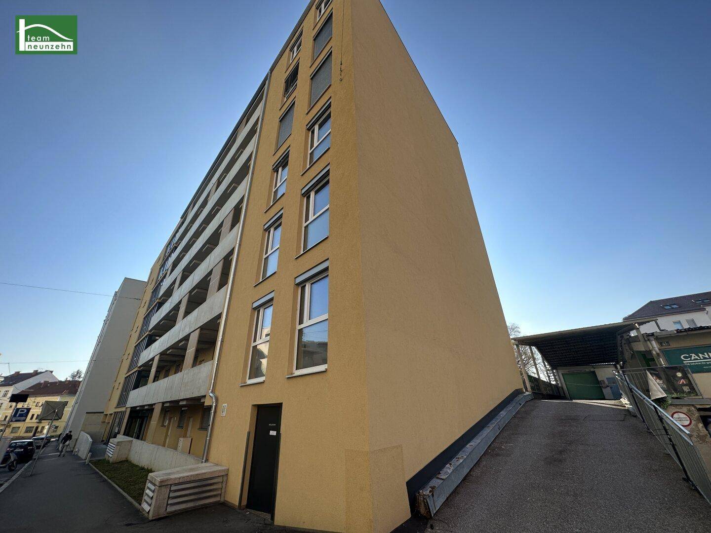 Seitenansicht eines modernen Wohngebäudes mit gelber Fassade und einer Zufahrtsrampe zur Tiefgarage.