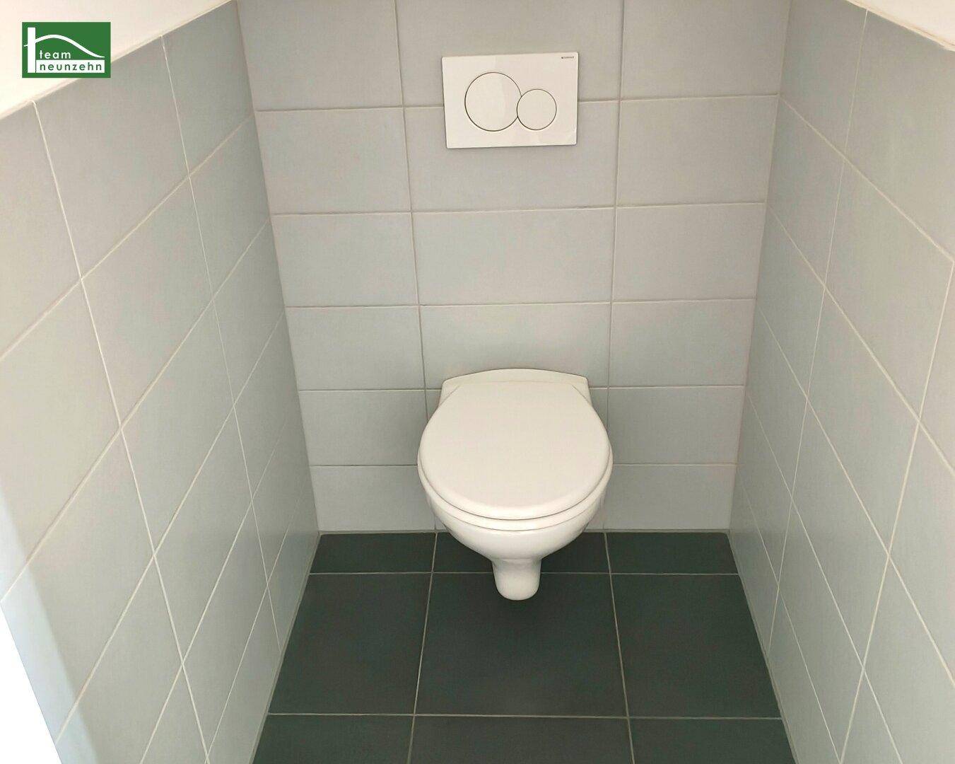 Separates WC mit wandhängender Toilette und hellgrauen Wand- und dunklen Bodenfliesen.
