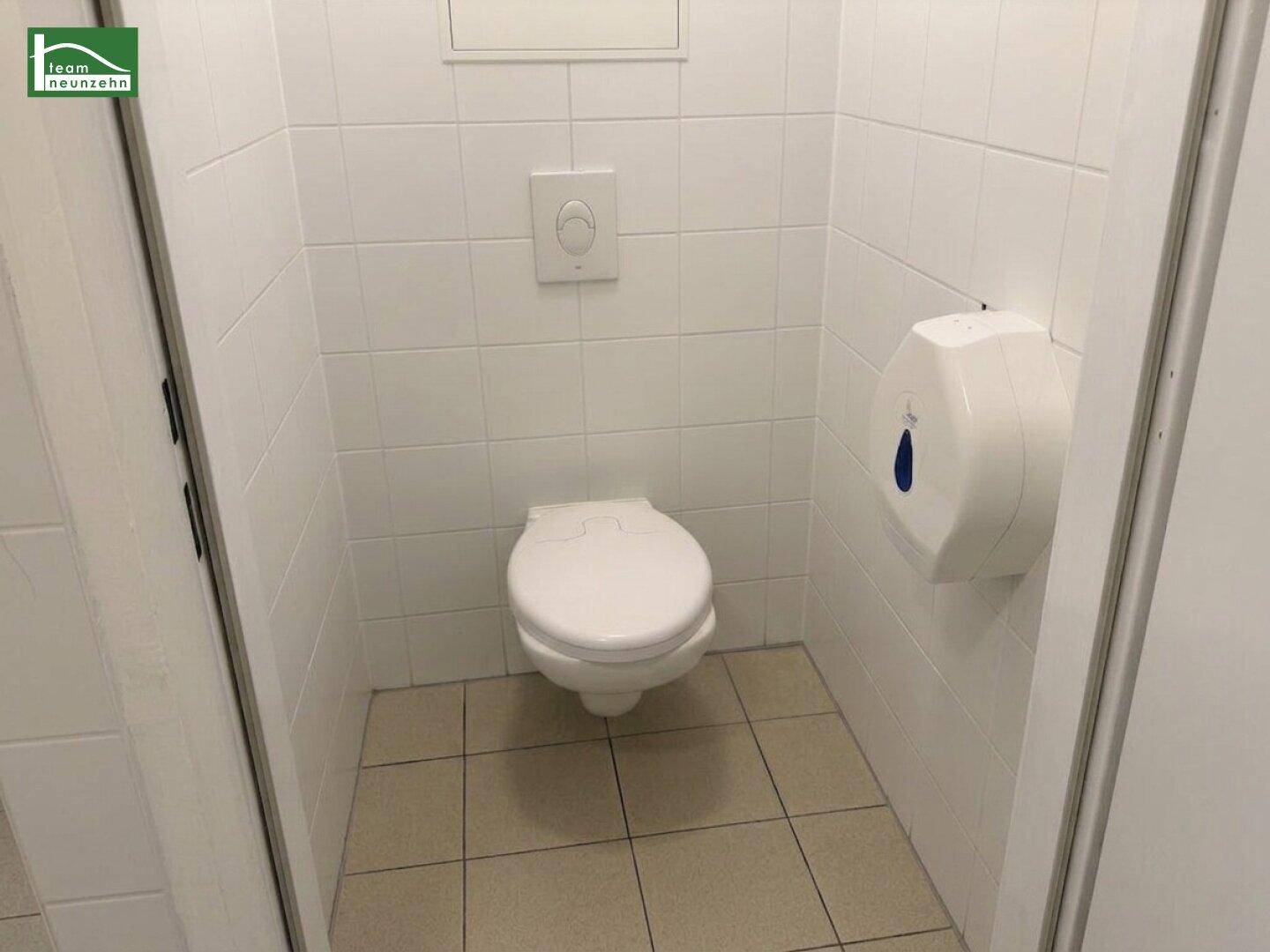 Einfache Toilette mit weißen Fliesen und Wand-WC, ausgestattet mit Papierhandtuchspender.