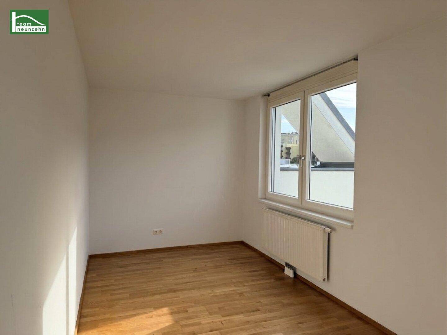 Helles Zimmer mit Holzboden und Fenster, das einen Blick auf die Dächer bietet.