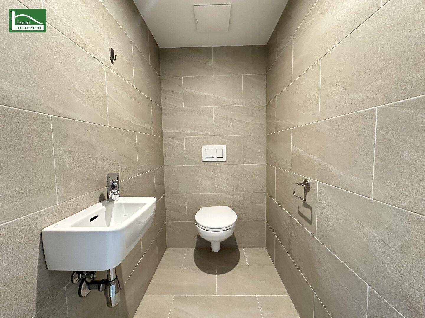 Modern gefliestes Gäste-WC mit wandhängender Toilette und kleinem Waschbecken.
