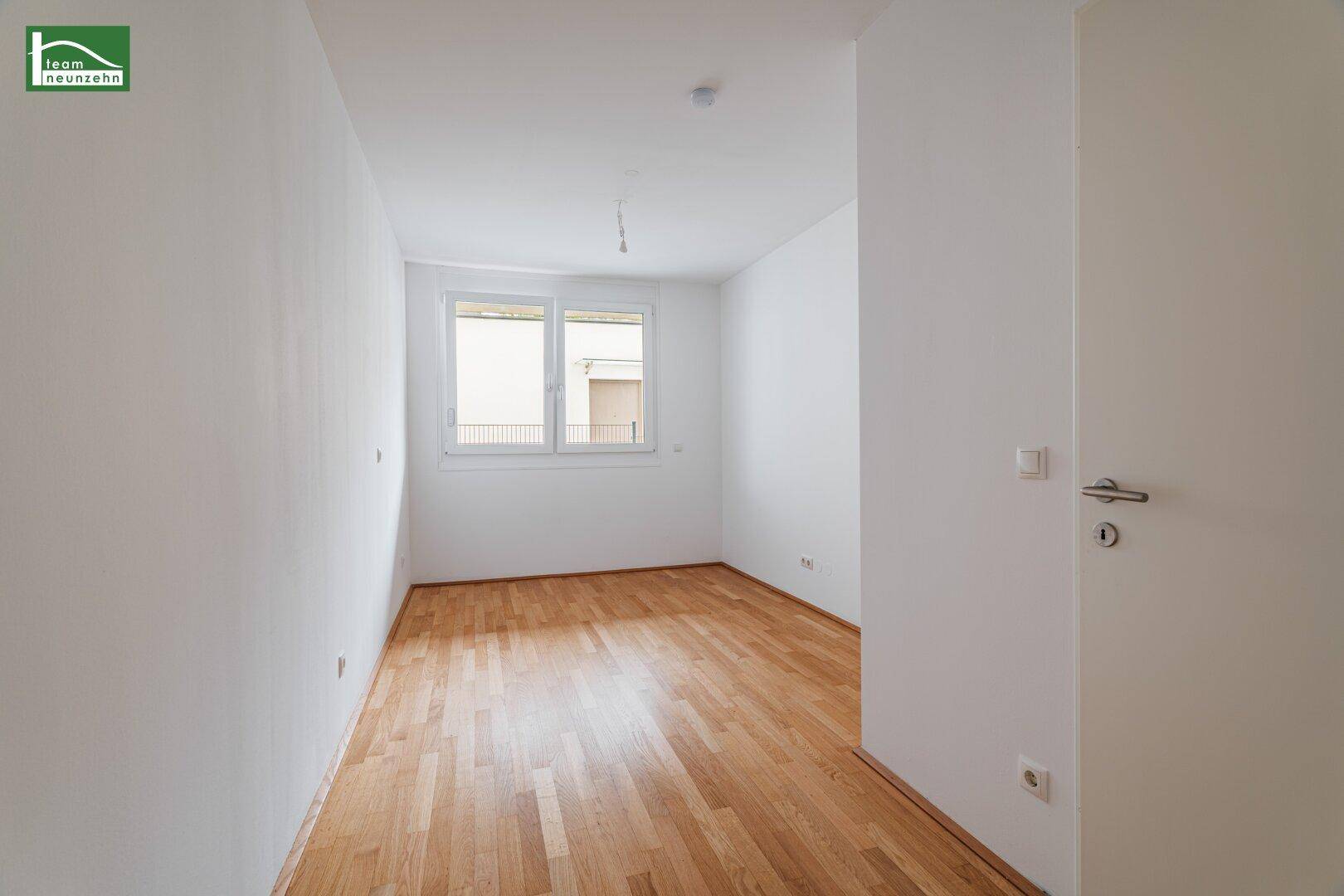 Helles Schlafzimmer mit Parkettboden und großem Fenster, das viel Tageslicht hereinlässt.
