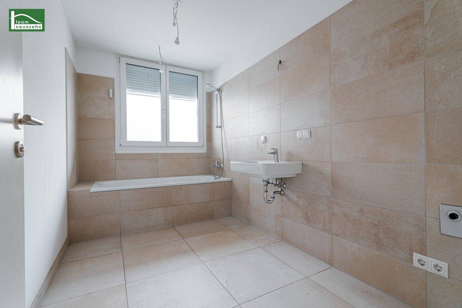 Modernes Badezimmer mit beigefarbenen Fliesen, Badewanne und Waschbecken unter einem Fenster.