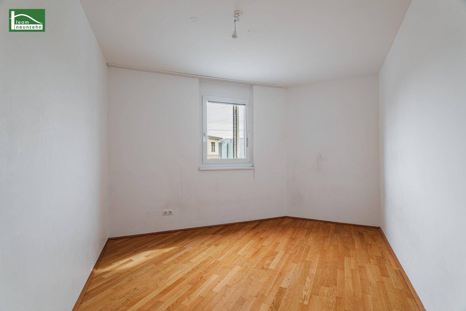 Heller, leerer Raum mit Parkettboden und einem Fenster, das Tageslicht hereinlässt.