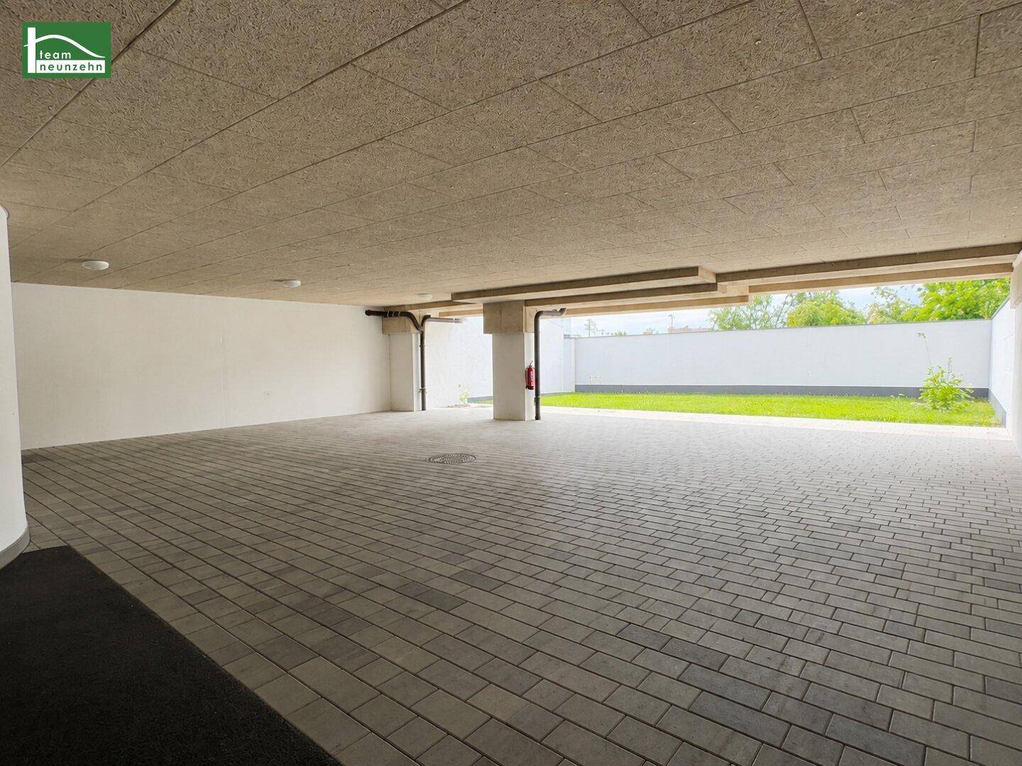 Überdachter Parkbereich mit gepflastertem Boden und Zugang zu einer Grünfläche.