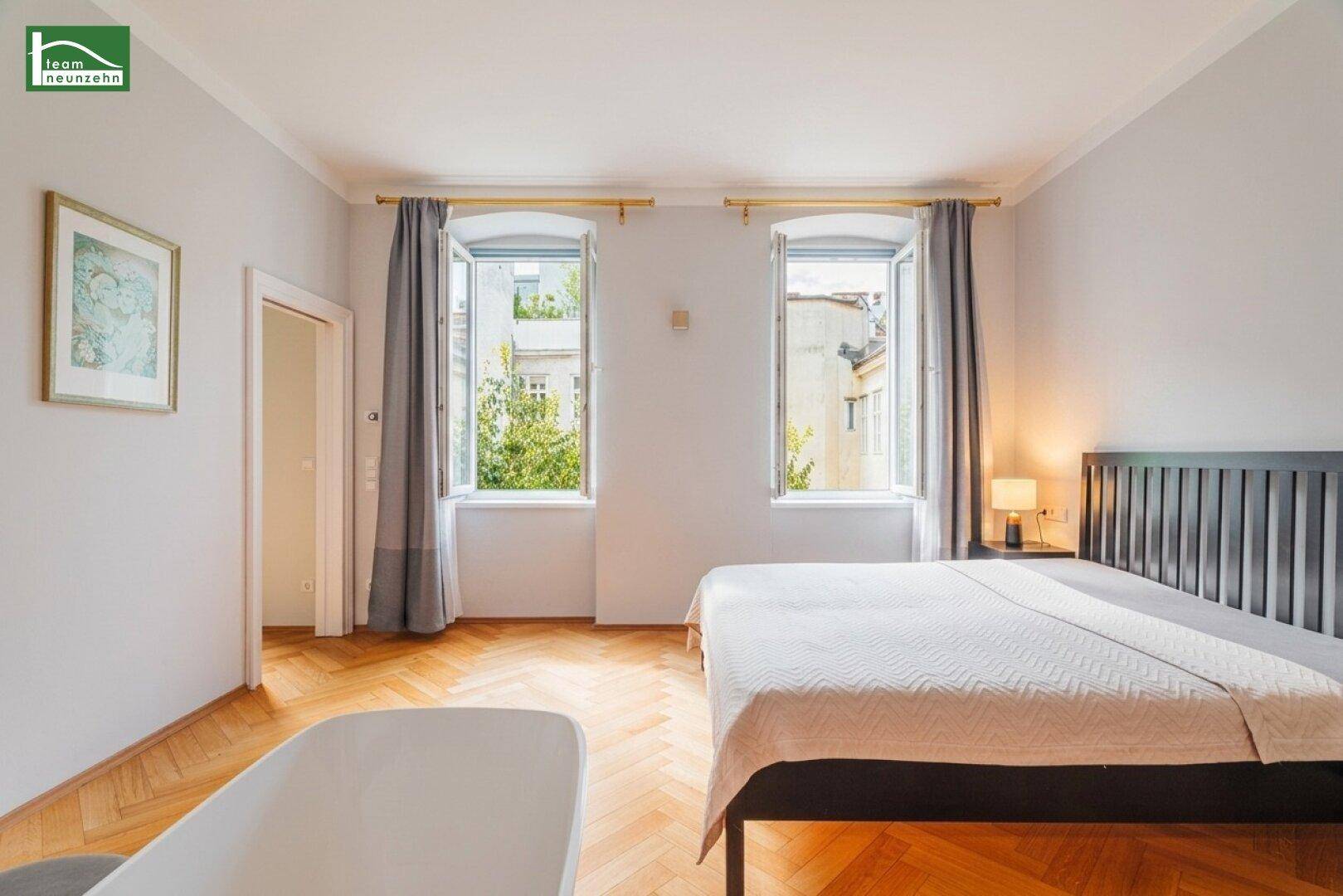 Helles Schlafzimmer mit Fischgrätparkett und zwei Fenstern mit Blick ins Grüne.