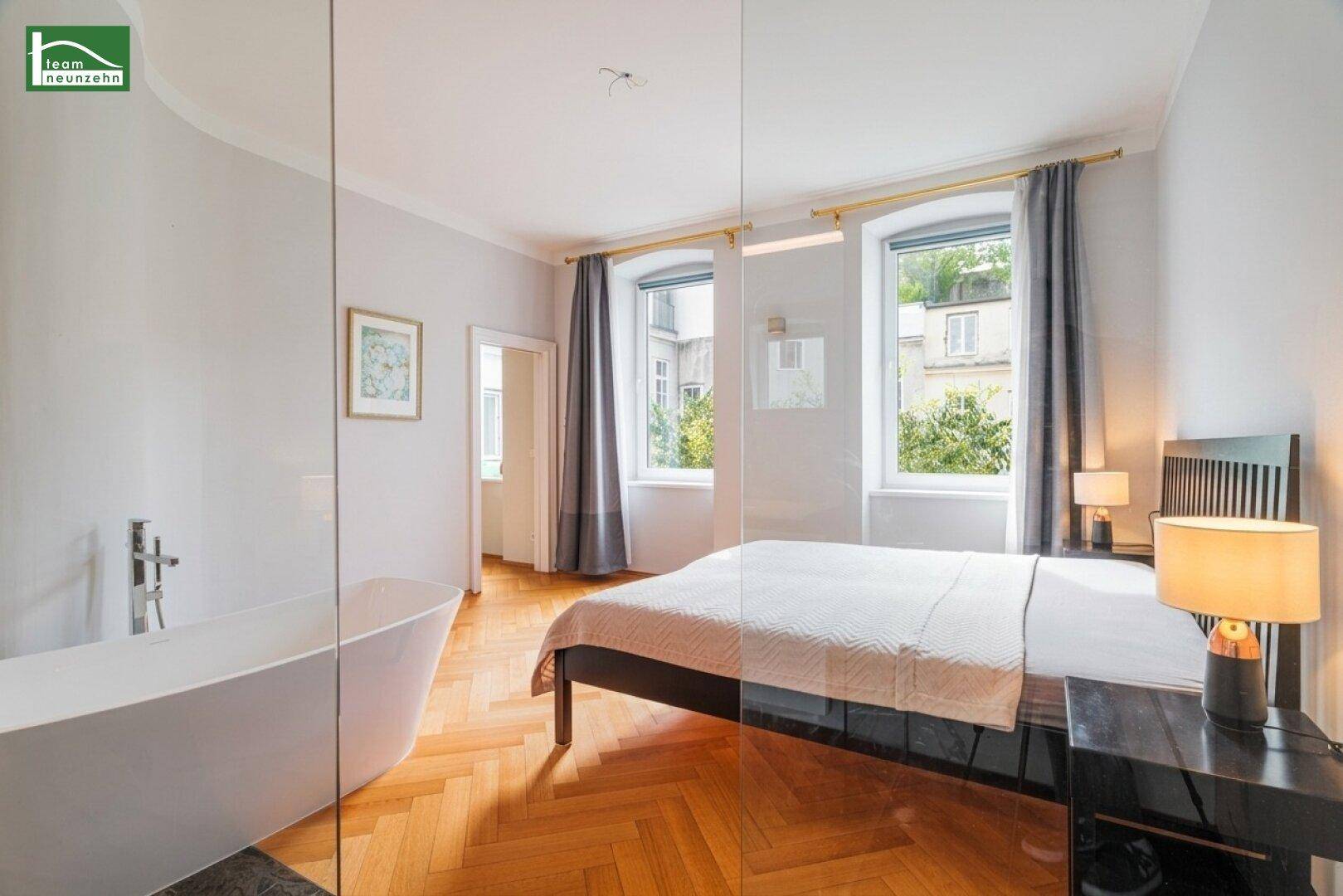 Schlafzimmer mit integriertem Badbereich, freistehender Badewanne und Blick aus dem Fenster.