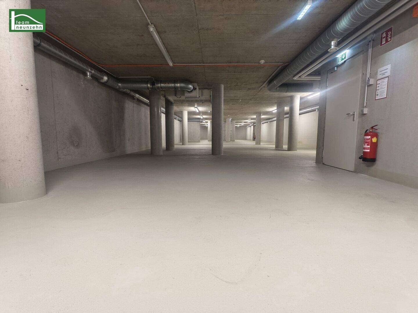 Langer Kellergang mit Betonpfeilern, glattem Boden und einer Brandschutztür am Ende des Raumes.