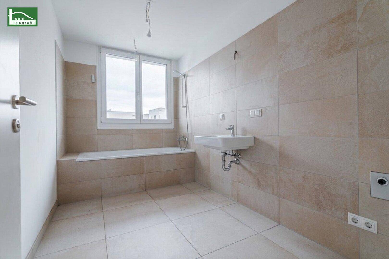 Helles Badezimmer mit beigefarbenen Fliesen, Badewanne, Waschbecken und großem Fenster für viel Tageslicht.