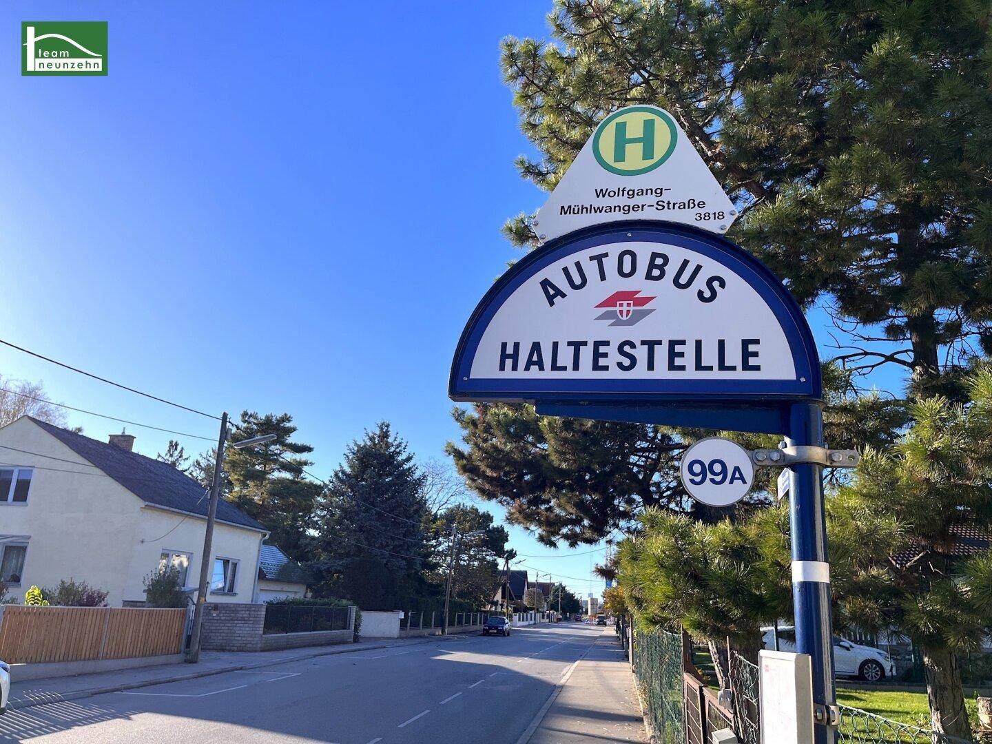 Bushaltestelle an einer ruhigen Wohnstraße mit Einfamilienhäusern und Bäumen unter blauem Himmel.