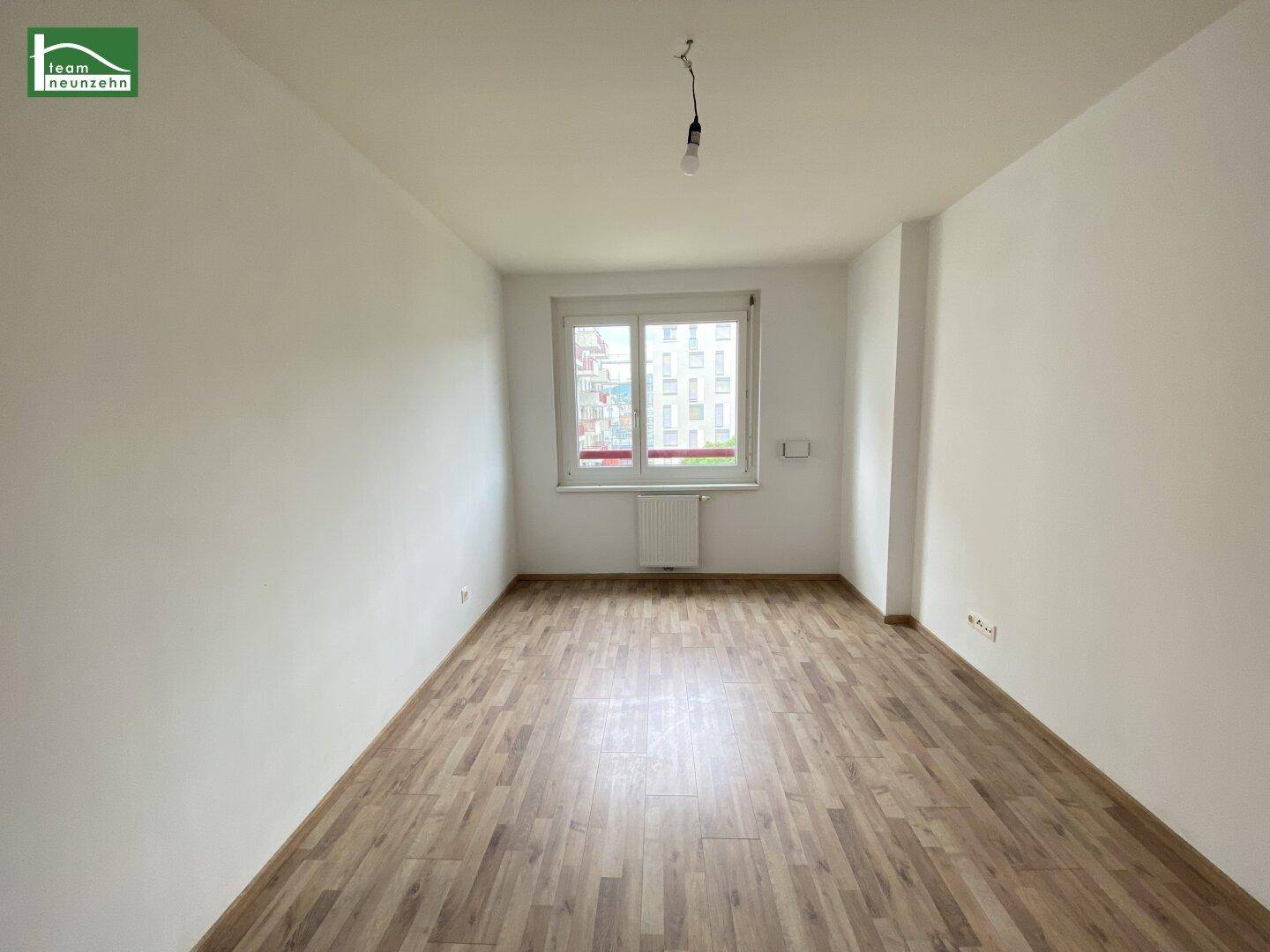 Leeres Schlafzimmer mit Laminatboden und einem Fenster, das viel Tageslicht hereinlässt.