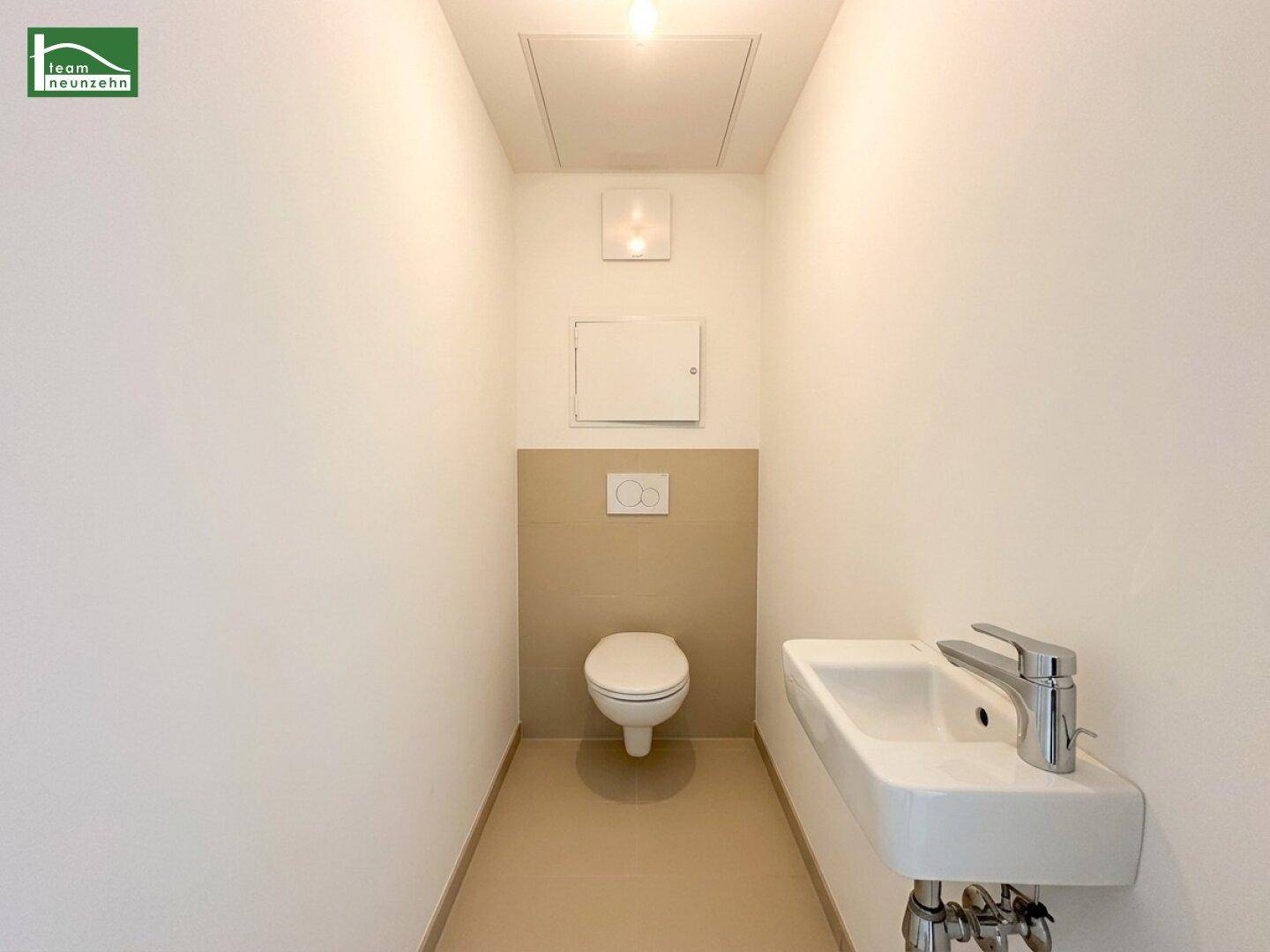 Separates WC mit wandhängender Toilette, kleinem Waschbecken und beigefarbenen Fliesen.