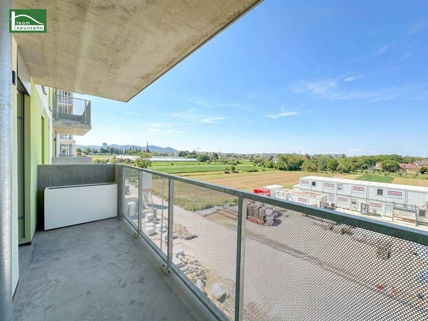 Geräumiger Balkon mit Glasgeländer und weitem Blick über grüne Felder und die Donau.