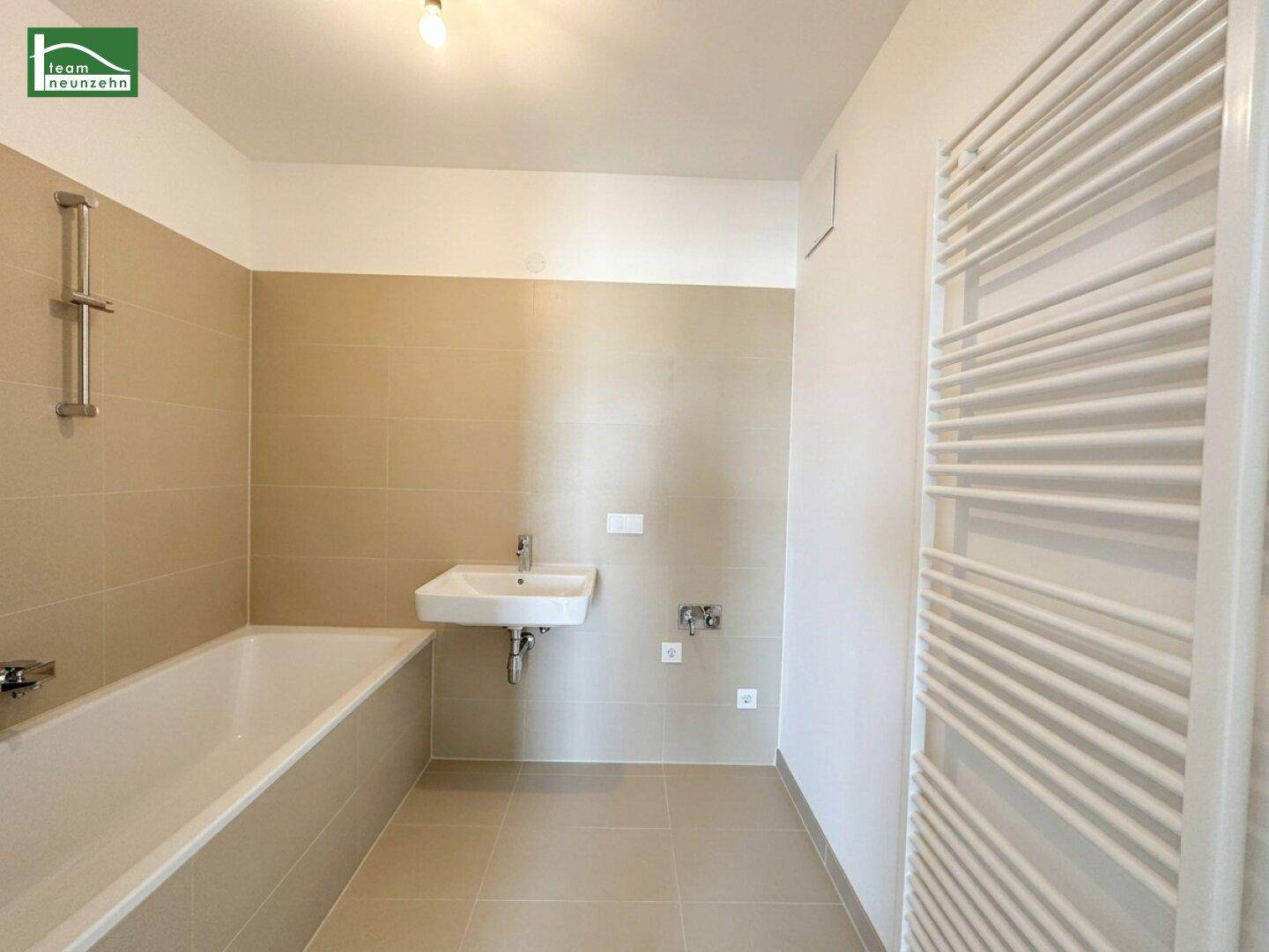 Modernes Badezimmer mit Badewanne, Einzelwaschbecken und beheiztem Handtuchhalter in beigen Fliesen.