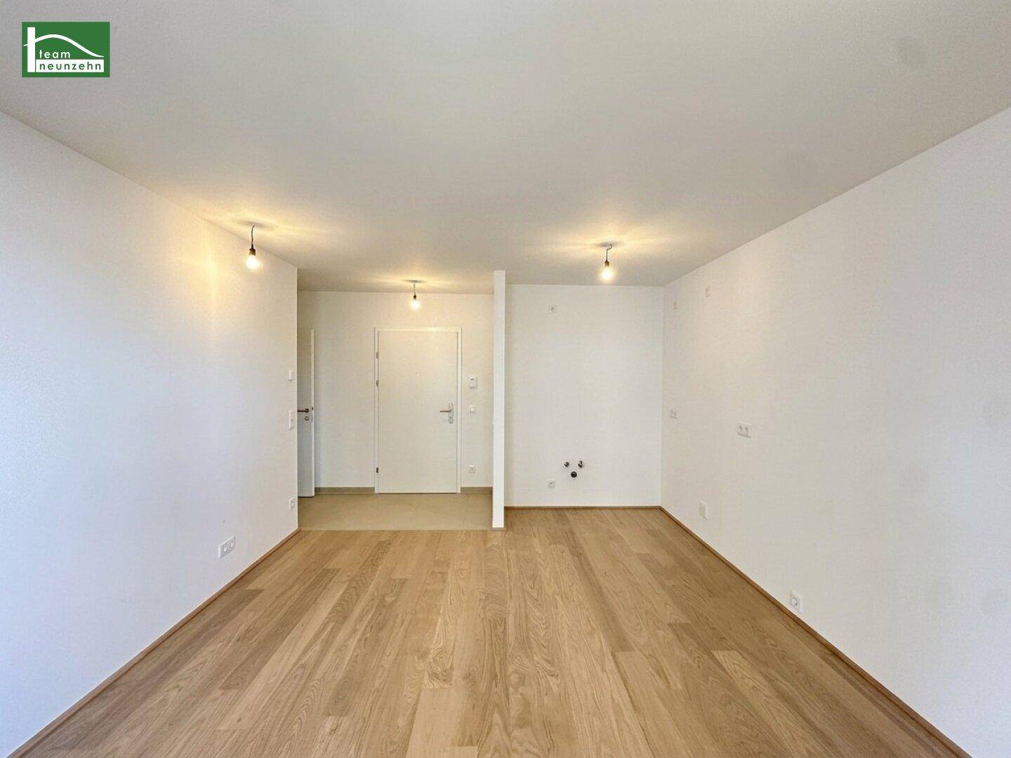 Geräumiges, leeres Zimmer mit Holzboden und weiß gestrichenen Wänden in einem Neubau.