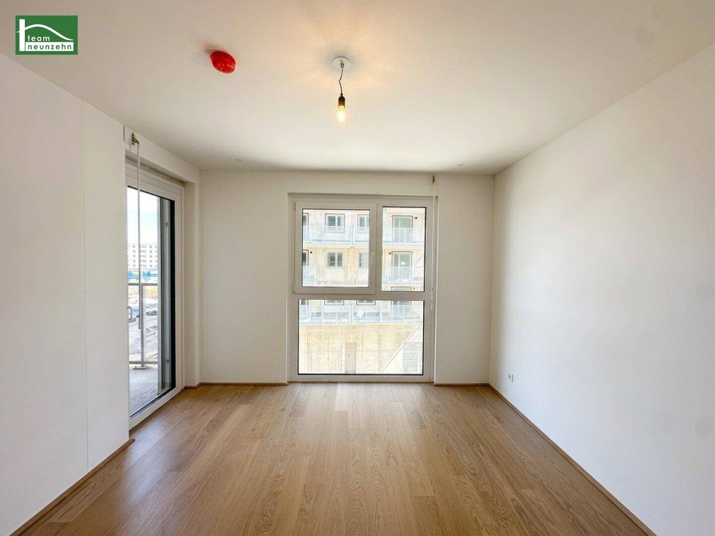 Helles, leeres Zimmer mit großem Fenster und Holzboden in einem Neubau.