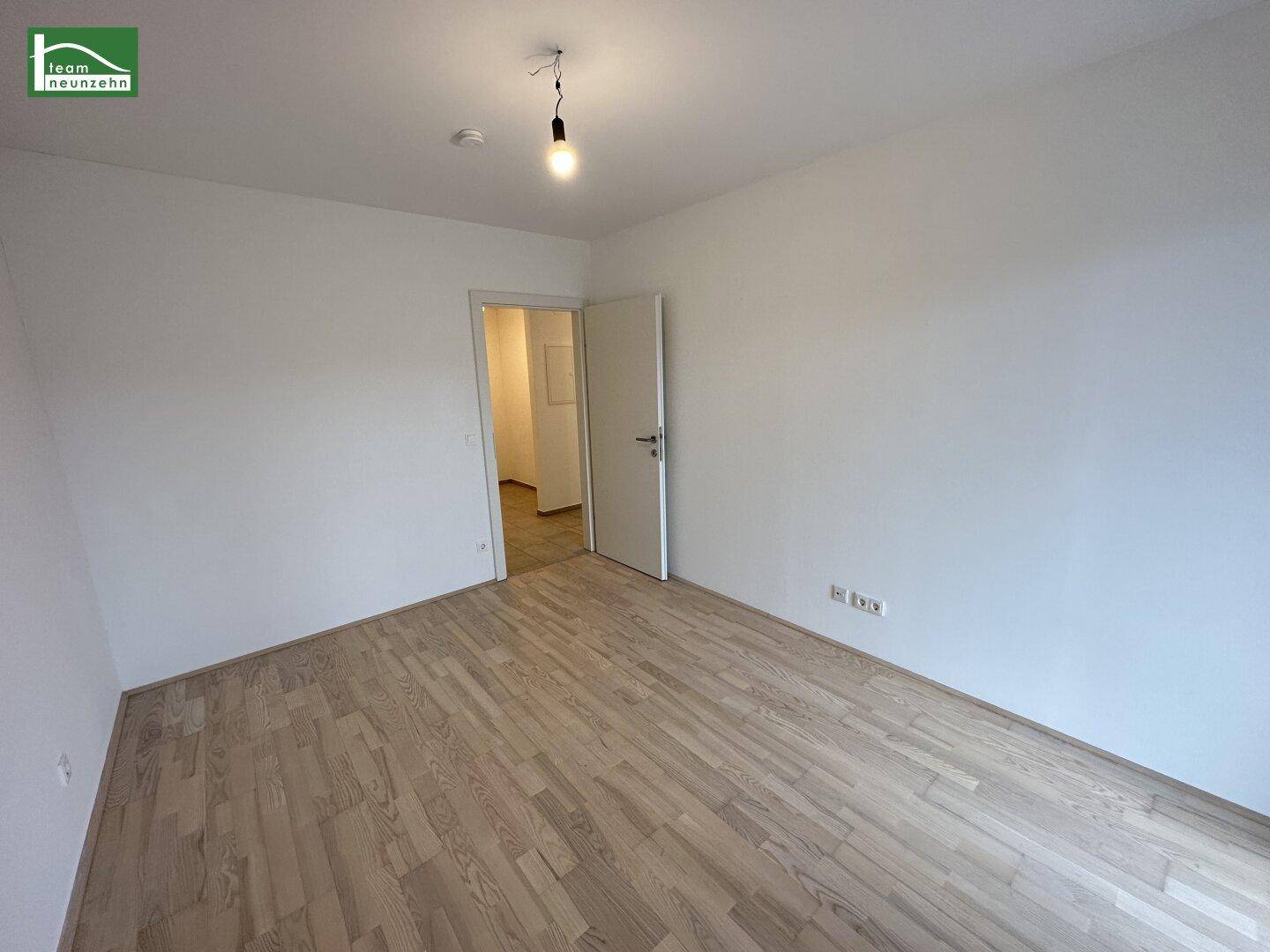 Helles, leeres Zimmer mit Holzboden und Zugang zu einem Flur.